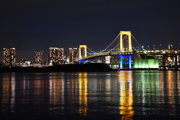 Fototapeta na wymiar The Rainbow Bridge is a suspension bridge in Odaiba, Tokyo. Illuminated city view with colorful reflection in the water, night view of the Japanese capital city with tall skyscrapers.