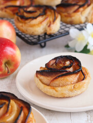 A baked mini caramelized apple rose tarts rolled in a rose form with puff pastry in a plate with its fresh fruit in background. 
