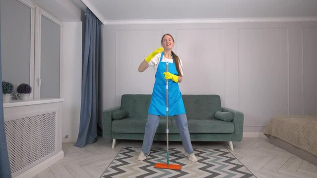 House cleaning with fun. Happy young housewife singing her favorite song while cleaning, using mop as microphone and guitar, enjoying her homework. Young woman dancing and cleaning in living room.