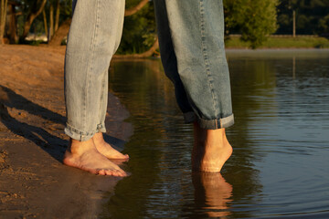Closeup of feet, legs of man and woman standing in water, female stands on her toes.Cheerful couple enjoys time together,having fun on beach. Green trees on background. Lovers wear denim. Horizontal
