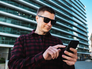 Young man in sunglasses using mobile phone in hands at modern new office building background.