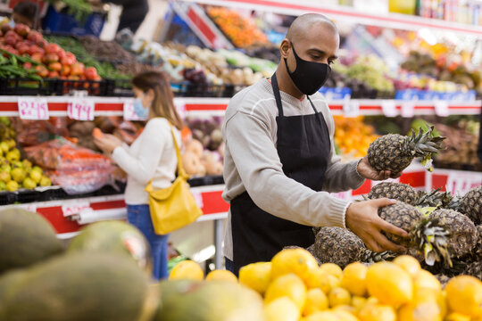 Latino-american Worker In Medical Mask In Supermarket With Pineapples During COVID-19 Pandemic