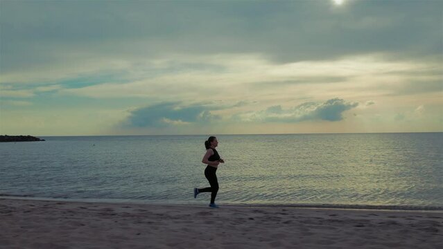 A young woman runs along the beach in sporty clothing, her hair blowing in the wind as she pushes herself to the limit.