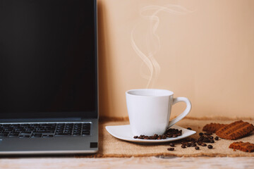 White cup of hot coffee with beans, cookies and laptop on beige table background