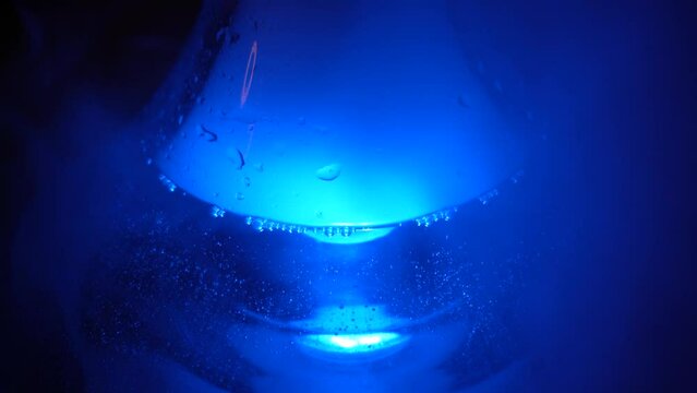 Close up of glass transparent flask of hookah on the table with a cloud of smoke. Shisha flask under the blue lamp light standing on a wooden table.