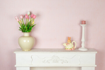 Spring easter home decoration with vase full of pink tulips flowers, bunny shaped porcelain jug and candle on white wooden rustic vintage fireplace and pink painted wall