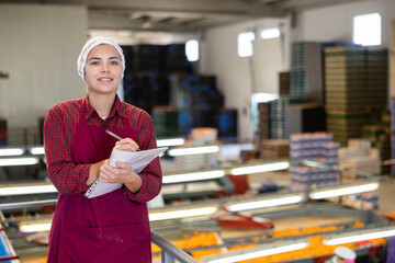 Positive young female supervisor standing with papers in hands while inspecting workflow of...