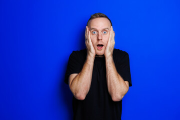 A young man in a black T-shirt on a blue background shows different emotions on his face. Cheerful man with a smile. Place for text. Male beauty