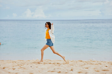 Fototapeta premium A woman runs along the beach in summer clothes on the sand in a yellow T-shirt and denim shorts white shirt flying hair ocean view