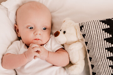 Little boy funny cute child baby lying on bed pillows with fluffy toy. Playful toddler with bulging eyes having fun, making faces grimaces. Happy childhood, family concept
