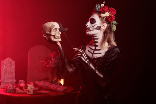 Glamour spooky woman acting creepy with skull in hand, holding black roses and wearing scary make up. Looking like santa muerte with flowers crown and body art on holy mexican celebration.