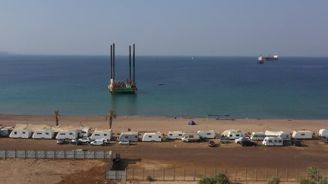 A jackup rig or a self-elevating unit mobile platform in an offshore area in front of Public HaDatiyim beach Camping site on Sunny day, Eilat City, Israel - aerial dolly right  