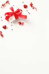 Happy Valentines Day flat lay composition. Gift box with red ribbon bow, confetti and party streamers on a white background. Vertical banner design.