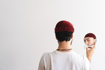 Young man with a raised eyebrow with his back turned wearing a brimless cap looking at camera through a mirror with a white background. Artistic photo. Copy space.