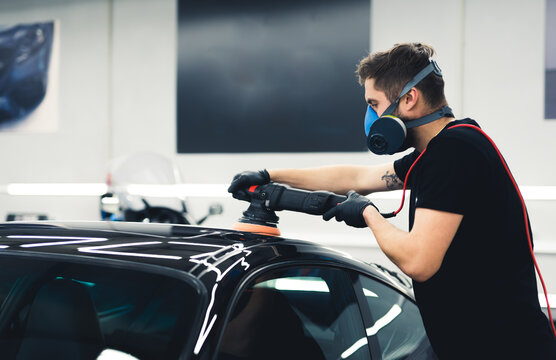 Caucasian man wearing protective face mask polishing black car paintwork using professional tool. Car detailing process. Horizontal indoor shot. High quality photo