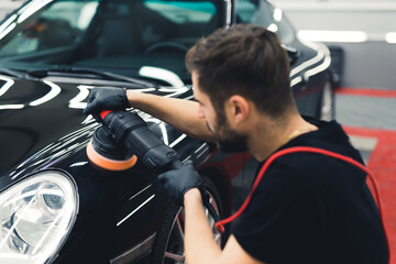 Fototapeta na wymiar Rear view of white man wearing black gloves squatting by the front of black car polishing it using professional buffer tool. Car detailing process. Horizontal indoor shot. High quality photo