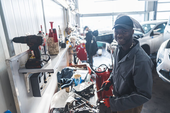 Medium Shot. A Black Mechanic Working By A Wokbench, Looking At The Camera And Smiling. Repair Shop Concept. High Quality Photo