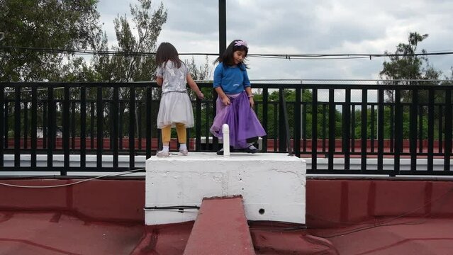 A couple of small latin girls playing in the rooftop of a building, they wear colorful dress. Red background
