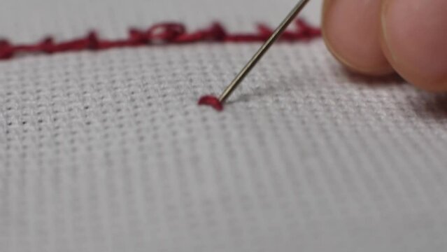 Close-up macro shot of a needle piercing the fabric while cross-stitching with red thread. Needlework, handmade jewelry and craft. The process of making an embroidered picture.