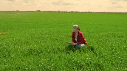 Naklejka na ściany i meble Working on farm with digital tablet in agriculture. Farmer woman with tablet evaluates with his hand shoots, green wheat sprouts in field. Technology of modern agriculture. Environmentally friendly