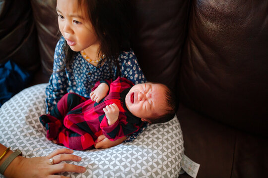 crying newborn in plaid pajama being held by toddler sister