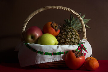 Wicker basket with a pineapple, some persimmons, tangerines and apples on a dark background and a red mat