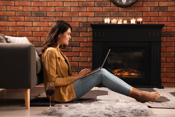 Fototapeta na wymiar Young woman with glass of wine and laptop near fireplace at home