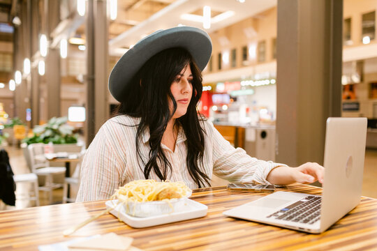 Woman eating noddle while using laptop in food court