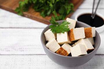 Bowl of smoked tofu cubes, soy sauce and parsley on white wooden table, closeup. Space for text