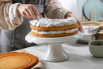 Woman smearing sponge cake with cream at white wooden table in kitchen, closeup