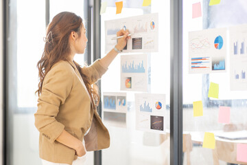 young asian entrepreneur of small company putting a adhesive sticky notes and graph chart in glass wall in office during analyzing strategy team meeting formulating business strategies.