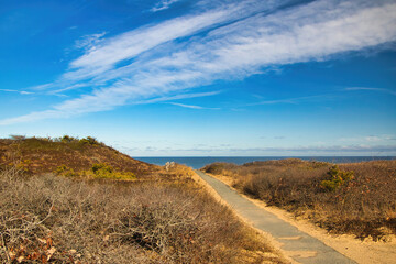 Under a partly sunny blue sky on a snowless Winter day, a trail passes through grassy sand dunes toward an ocean horizon on Cape Cod, MA,