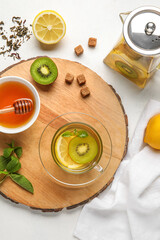 Board with glass cup of fruit tea, kiwi, lemon, mint, honey and teapot on white background