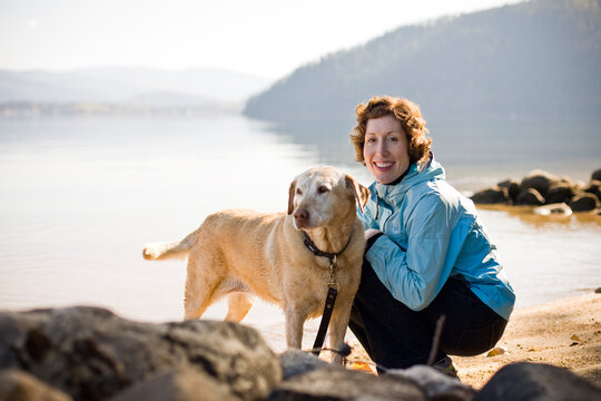 A smiling young woman with red hair and a blue jacket sits next to a golden labrador on the shores of a lake.