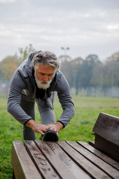 Senior Male Stretching Leg On Wooden Bench While Exercising At Park 