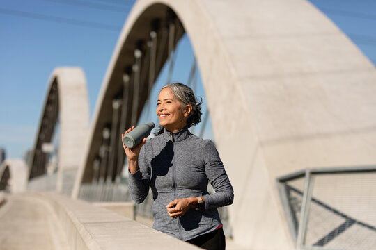 Smiling Woman Holds A Water Bottle On A Morning Run On A Bridge - Powered by Adobe