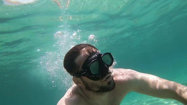 Front view of a man diving and blowing bubbles from his mouth in slow motion in a turquoise sea.