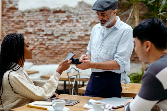 Woman in cafe interacting with waiter while paying with mobile phone