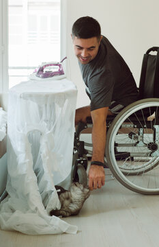 A kitten distracts a man in a wheelchair while he irons the curtains.
