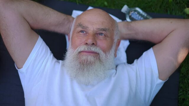 Top view happy senior Caucasian man lying on exercise mat smiling resting after workout outdoors. handsome bearded grey-haired old sportsman enjoying healthy lifestyle in summer park