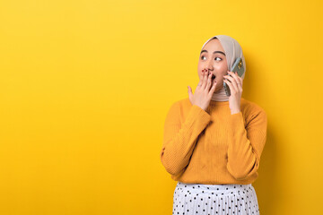 Beautiful amazed Asian girl wearing hijab talking on cell phone with friend, looking at empty space isolated on yellow background - obrazy, fototapety, plakaty
