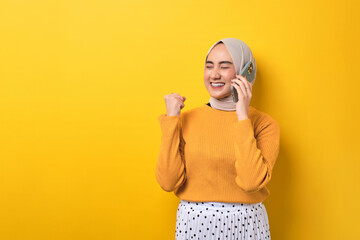 Beautiful excited Asian girl wearing hijab talking on cell phone, getting good news, raising fist,...