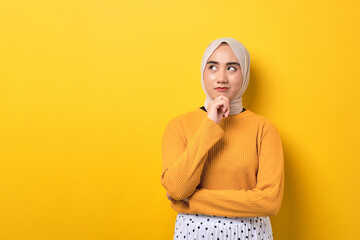 Beautiful pensive Asian girl wearing hijab looking aside at empty space, touching chin, thinking about offer isolated on yellow background