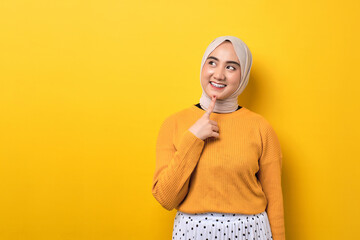 Beautiful thoughtful Asian girl wearing hijab looking into empty space, touching chin, thinking about something interesting isolated on yellow background - obrazy, fototapety, plakaty