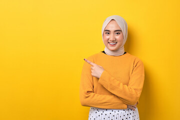 Beautiful cheerful Asian girl wearing hijab pointing finger at empty copy space isolated on yellow background