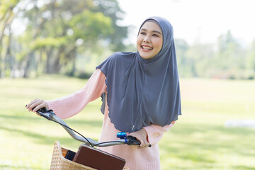 Smiling Asian young Muslim woman wear hijab headscarf and hijab dress walking with her bicycle and books in the park