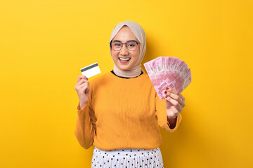 Beautiful cheerful Asian girl wearing hijab showing money banknotes and credit card isolated on...