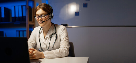 Attractive doctor wears a headset use laptop webcam for video call or conference, working at night shift. Remote online medical chat consultation, virtual physician. Telemedicine
