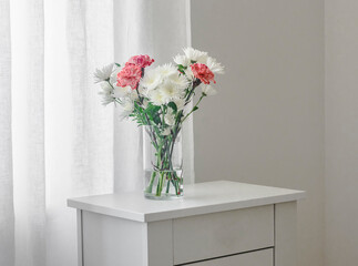 Fresh chrysanthemums and carnations in a glass vase on a white chest of drawers in a cozy bright room