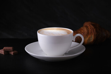 Cappuccino with croissant on a black background. White cup with cappuccino on a dark background. Italian breakfast on a black background.